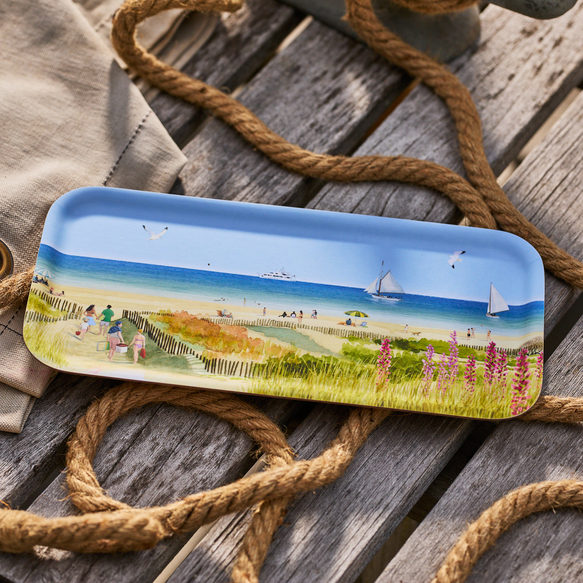 The Caskata X Felix Doolittle Beach Dunes Small Rectangular Tray features a painted beach scene with people, sailboats, seagulls, and flowers—ideal summer decor from Caskata. It rests on wood beside rope and a beige cloth.