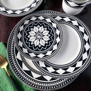 A set of sophisticated dinnerware with Caskata Artisanal Home's Fez Rimmed Dinner Plate, featuring black and white geometric patterned plates and bowls arranged on a dark brown table, accompanied by a green napkin and a fork.