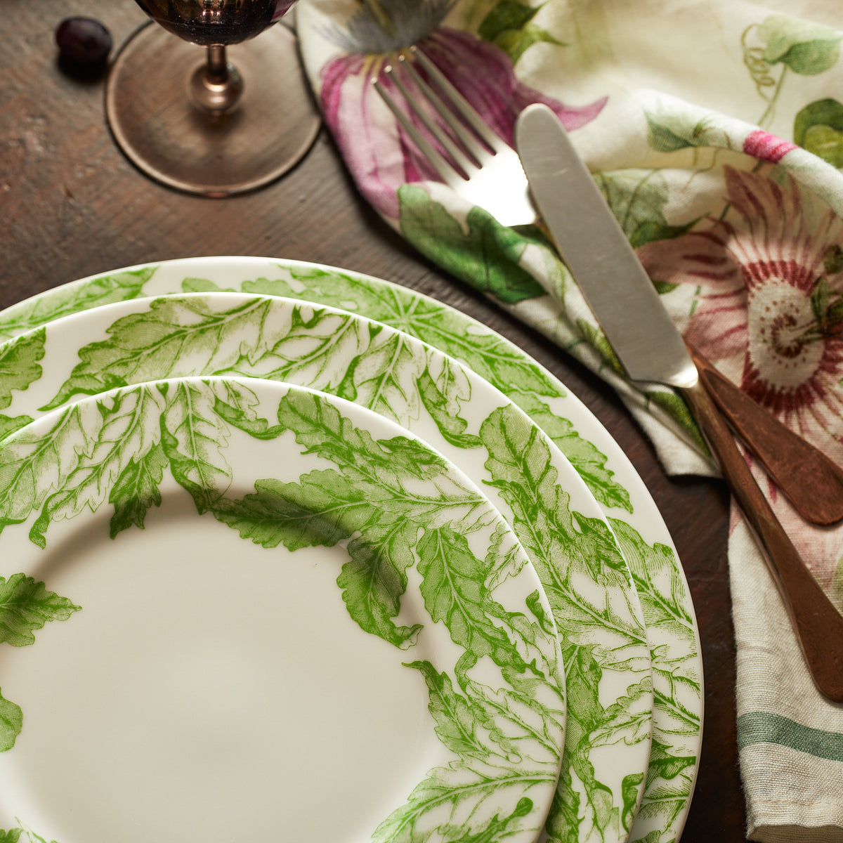 A set of three Freya Rimmed Charger Plates from Caskata Artisanal Home, with green leaf patterns, reminiscent of green florals, stacked on a wooden table, accompanied by a knife, fork, and floral napkin. A partial view of a wine glass is visible in the top left corner.