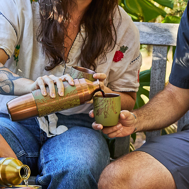 A person pours a drink from a Caskata x High Camp Freya Parkside Flask by Caskata into a cup held by someone else while they sit outdoors on a bench.