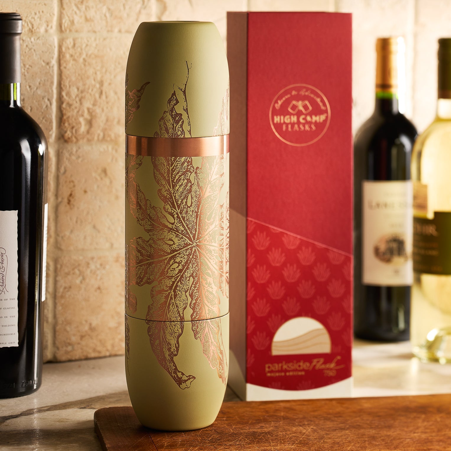 A Caskata x High Camp Freya Parkside Flask with a leaf design stands beside its red and white box, with wine bottles in the background on a kitchen counter.