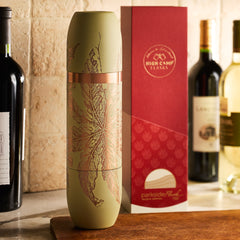 A Caskata x High Camp Freya Parkside Flask with a leaf design stands beside its red and white box, with wine bottles in the background on a kitchen counter.