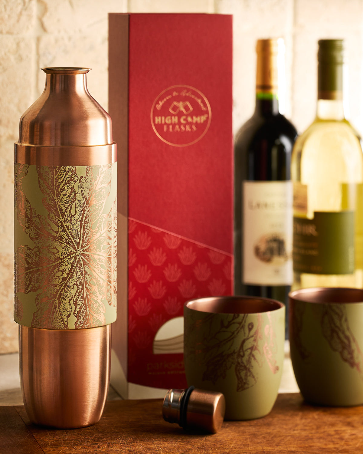 The Caskata x High Camp Freya Parkside Flask by Caskata, shown with a copper cocktail shaker set, two matching cups, and a red gift box beside wine bottles on the countertop.