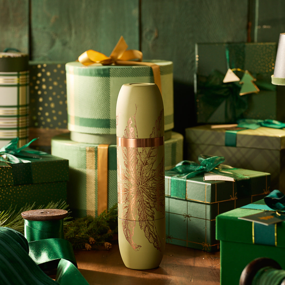 A tall, green cylindrical gift box with a leaf design stands on a table beside green-wrapped presents, ribbons, and the Caskata x High Camp Freya Parkside Flask by Caskata.