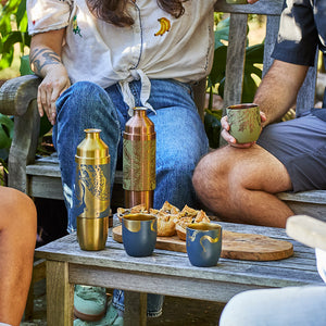 Three people sit outdoors on a wooden bench with drinks in decorated cups, the Caskata x High Camp Dark Blue Lucy Parkside Flask by Caskata, and a wooden tray of snacks in the center.