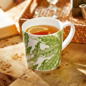 A Caskata Freya Ultimate Set white porcelain mug with green leaf patterns, filled with tea and lemon, sits on a wooden table next to a wicker tray. Dishwasher- and microwave-safe for convenience.