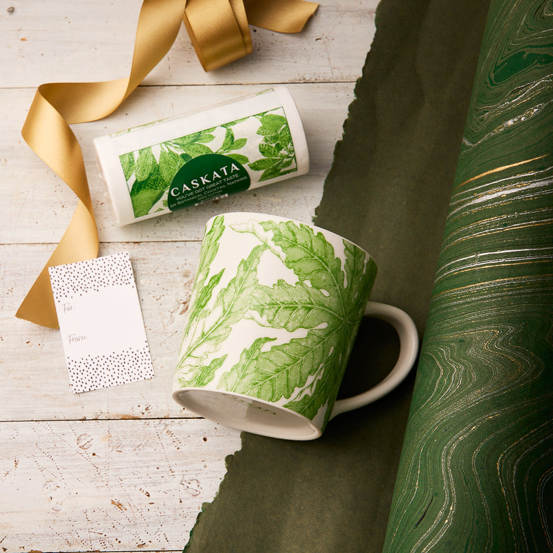 The "Freya for a Friend" Bundle by Caskata features a green and white fern-patterned porcelain mug with matching packaging, gift tag, gold ribbon, and green marbled wrapping—creating a charming gift set on a white wooden surface.
