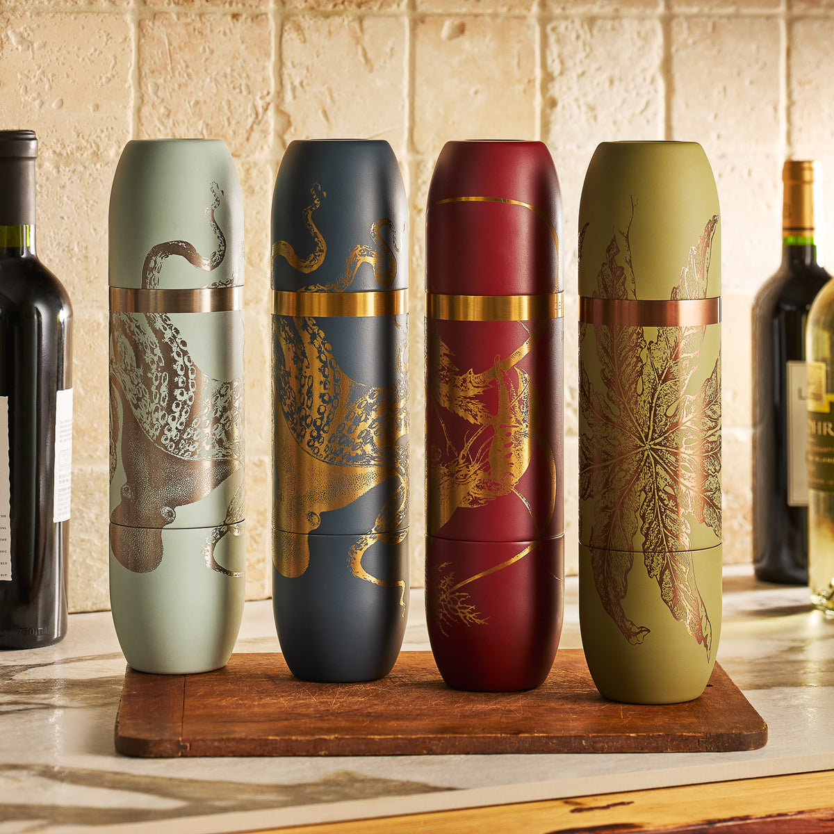 Four decorative spice grinders with gold botanical and animal designs stand on a wooden board, next to the Caskata x High Camp Dark Blue Lucy Parkside Flask by Caskata, with wine bottles set against a tiled backsplash.
