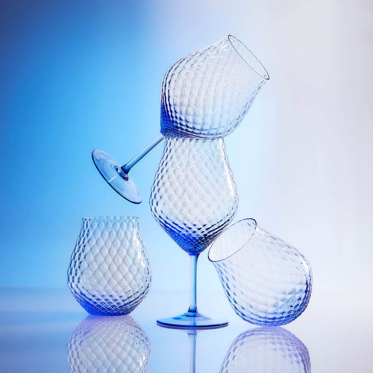 Four textured, blue-tinted glasses, three without stems and one wine glass with a stem, are artistically stacked against a gradient blue background. Made from lead-free crystal in the Czech Republic, these elegant Phoebe Cobalt Universal Wine Glasses from Caskata add a touch of sophistication to any setting.