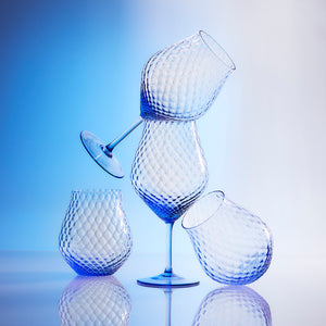 Four textured, blue-tinted glasses, three without stems and one wine glass with a stem, are artistically stacked against a gradient blue background. Made from lead-free crystal in the Czech Republic, these elegant Phoebe Cobalt Universal Wine Glasses from Caskata add a touch of sophistication to any setting.