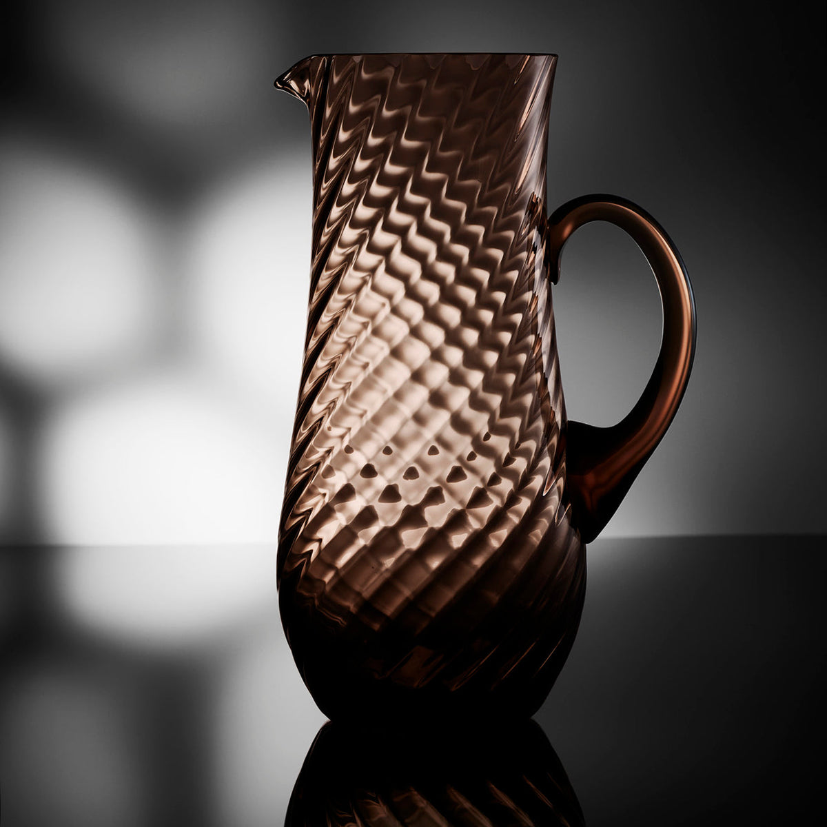 The Quinn Mocha Pitcher is backlit to showcase it&#39;s beautiful swirled brown glass. This handled piece from Caskata is delicate to look at yet substantial in feel.
