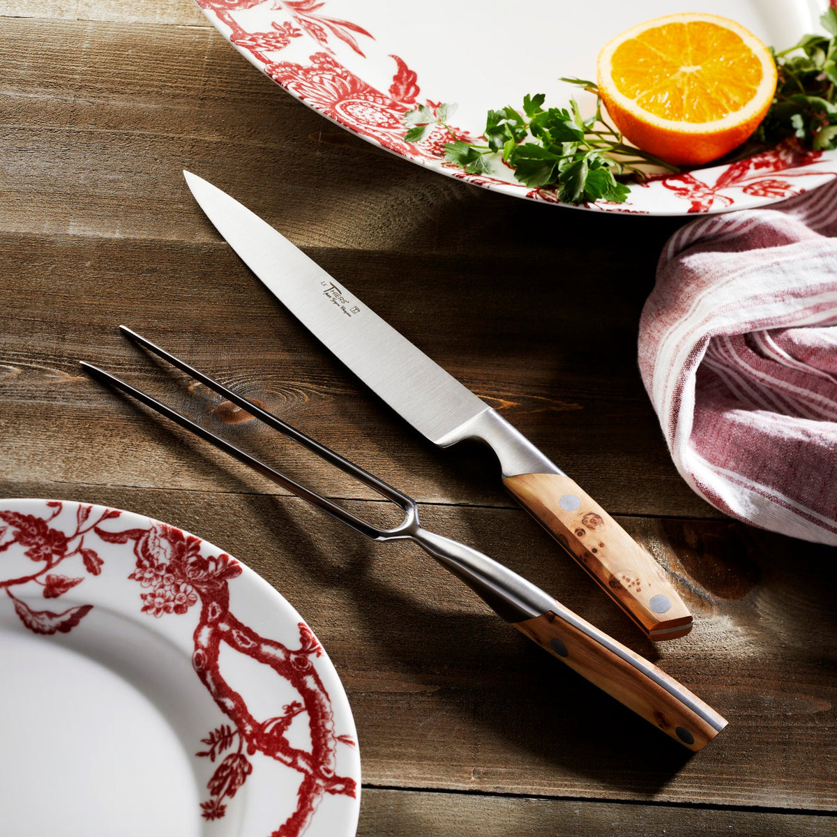 A Goyon-Chazeau Juniper Wood Carving Set with a knife and fork on accompanied by Caskata&#39;s Arcadia Crimson dinnerware..