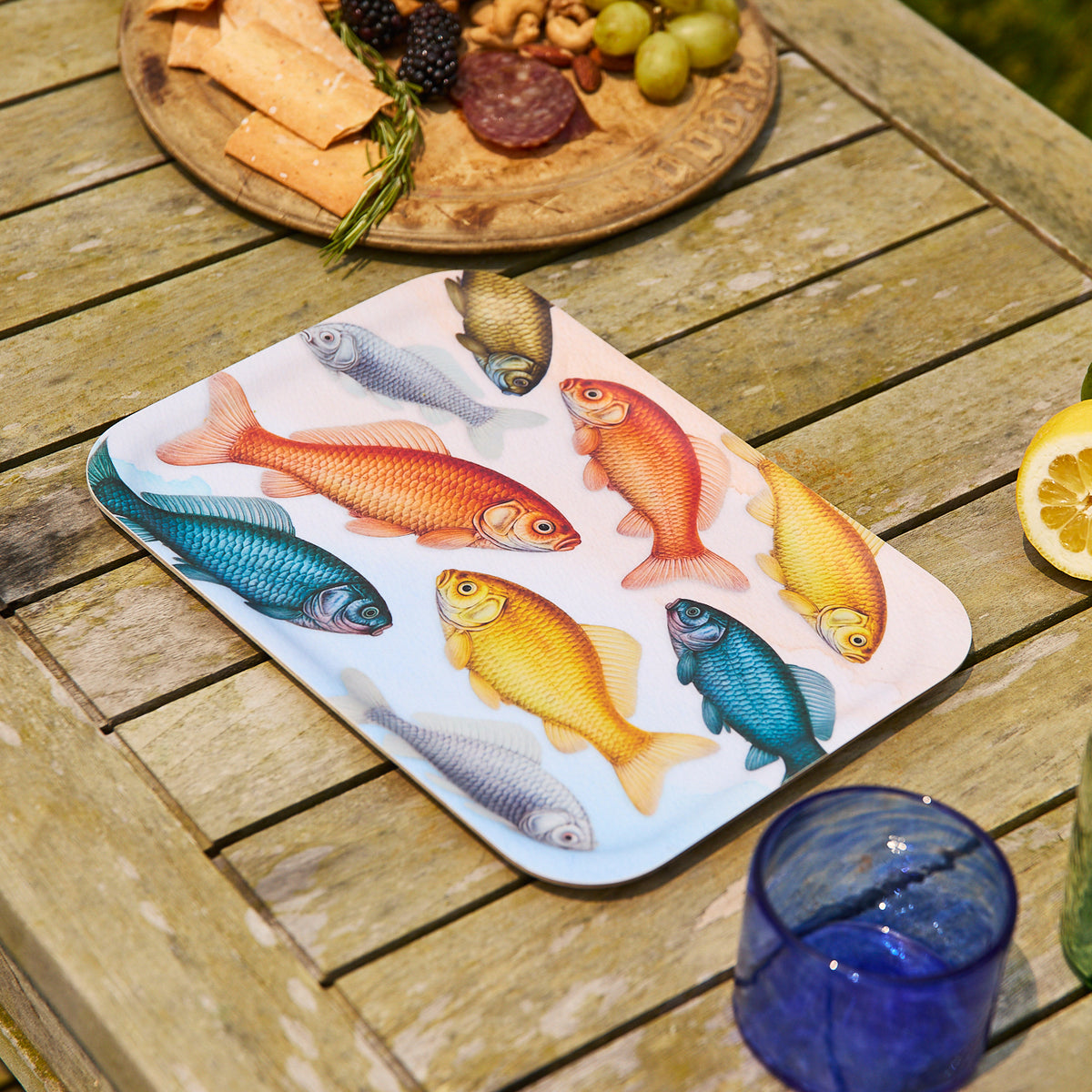 The Caskata Goldfish Medium Rectangular Birchwood Tray, styled with a blue glass, lemon slice, and charcuterie plate with crackers, grapes, and berries—features colorful fish art and is made in Finland.