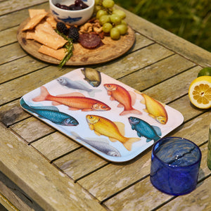 The Caskata Goldfish Medium Rectangular Birchwood Tray, made in Finland, features colorful fish illustrations and sits on a wooden table beside snacks, fruit, a blue glass drink, and sliced lemon.