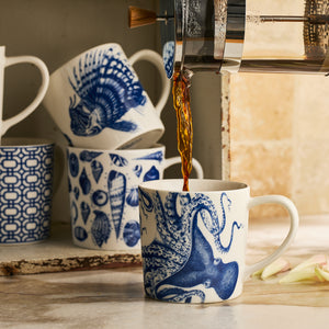Coffee is being poured into a Lucy Mug from Caskata Artisanal Home, its blue and white porcelain design standing out as other matching mugs are stacked in the background.