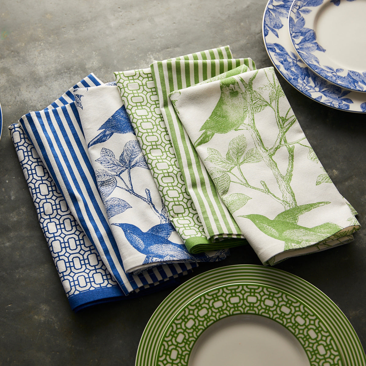 The Caskata Arbor Birds Verde Dinner Napkins, made of 100% cotton and featuring blue and green arbor patterns, are elegantly placed beside matching white plates on a gray surface. Machine washable, these napkins combine style with practicality.