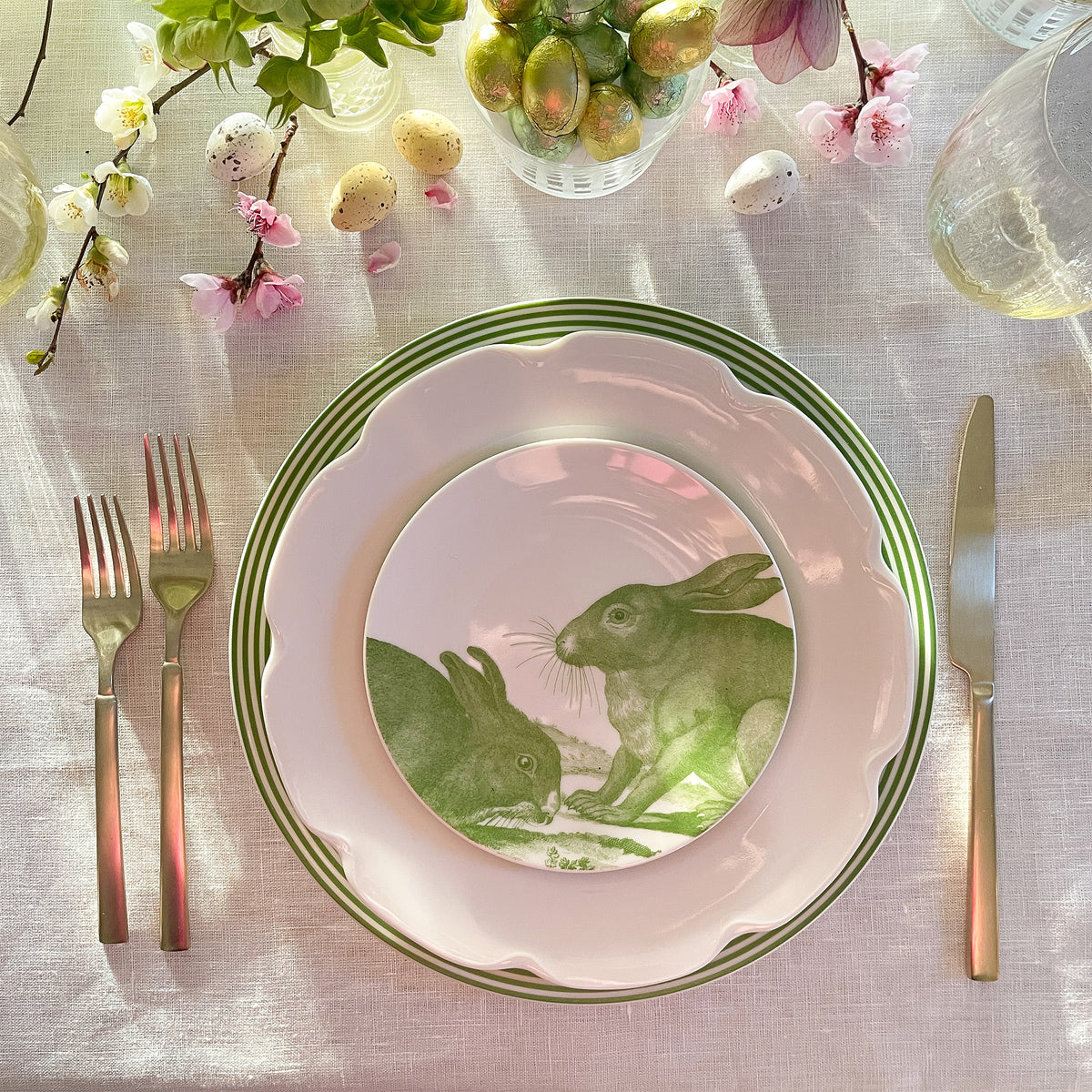 A table setting with a white plate featuring a charming bunny scene, surrounded by Caskata Bunnies Verde Small Plates and canapé plates. Silver cutlery, decorated with flowers, Easter eggs, and glasses complete the look on a white tablecloth. The high-fired porcelain adds an elegant touch to the festive arrangement.