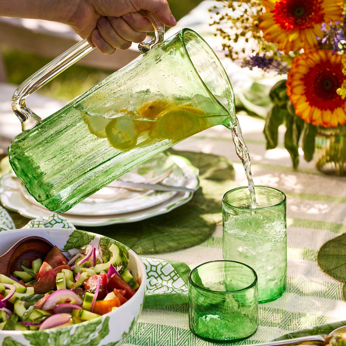 A hand pours lemon water from a green recycled glass pitcher into a Caskata Viento Small Green Tumbler, part of a set of 6, on an outdoor table with salad, plates, and flowers.