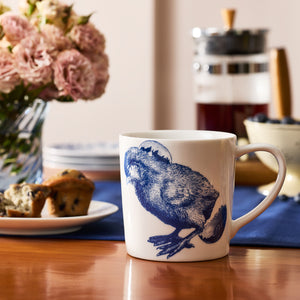 The Grumpy Chick Mug by Caskata, featuring a blue chicken illustration, rests on a wooden table next to a blueberry muffin, fresh flowers, and a French press—a delightful porcelain drinkware choice for your morning routine.