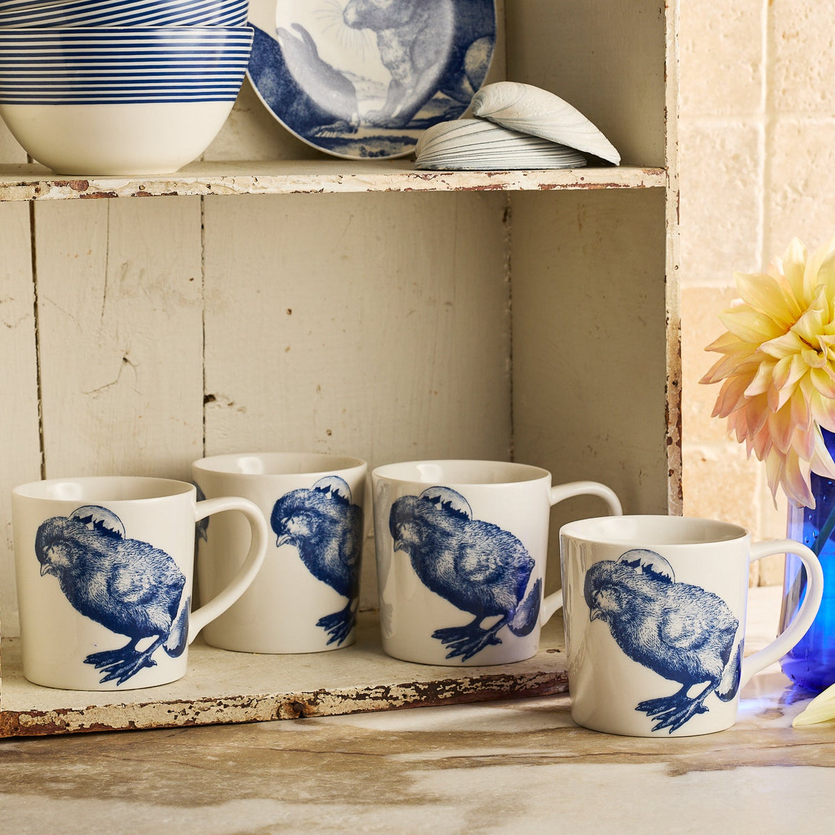 Four Caskata Grumpy Chick Mugs, white with blue chicken illustrations, are displayed on a rustic kitchen shelf with other blue and white ceramics.