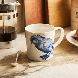 The Caskata Grumpy Chick Mug, a blue botanical cup with a quirky chicken design, sits on a countertop alongside a spoon, French press, and plate of food.