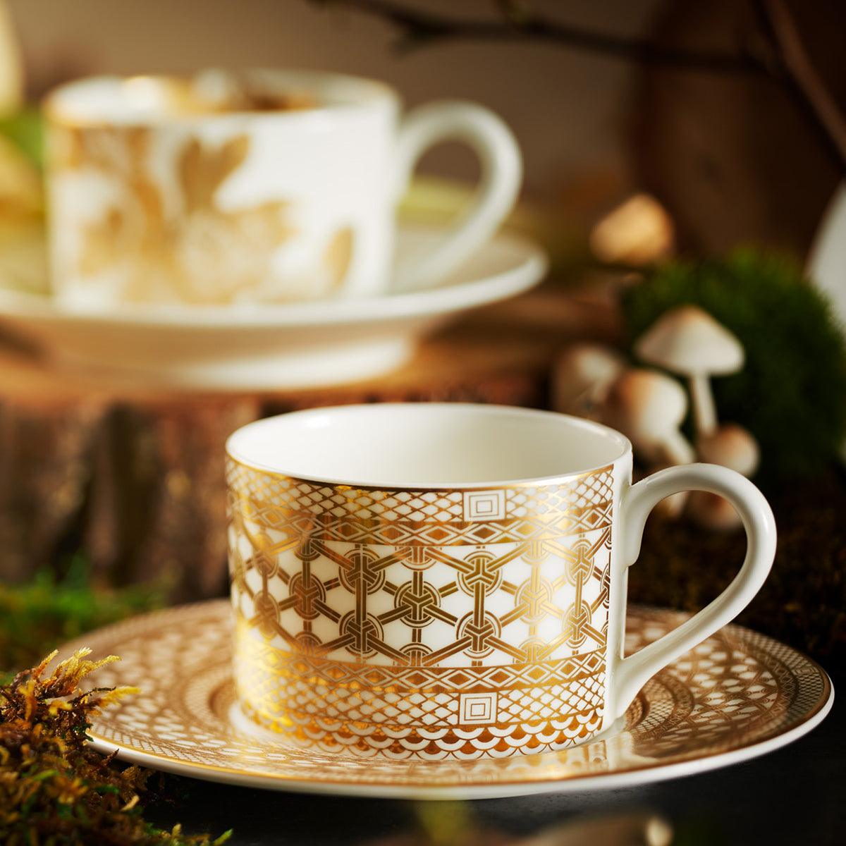 Close-up of two Hawthorne Gilt tea cups and saucers from the Caskata Artisanal Home collection, showcasing intricate golden patterns and surrounded by greenery and mushrooms.