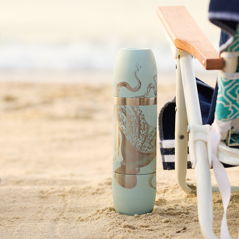The Caskata x High Camp Light Blue Lucy Parkside Flask, featuring an octopus design and crafted from Sommelier-Grade stainless steel, stands upright on sand beside a beach chair draped with a towel.