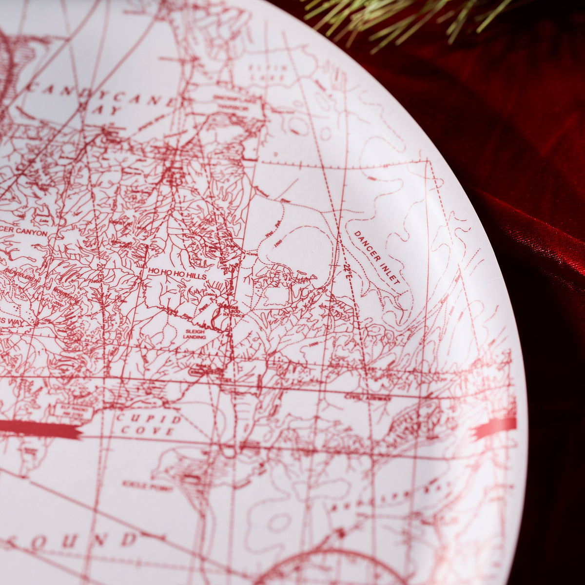 Close-up of Caskata&#39;s White Holiday Hunt Large Round Tray, featuring a detailed red map design with labeled areas like &quot;Danger Isle&quot; and &quot;Cupid Cove,&quot; displayed on a red fabric background.