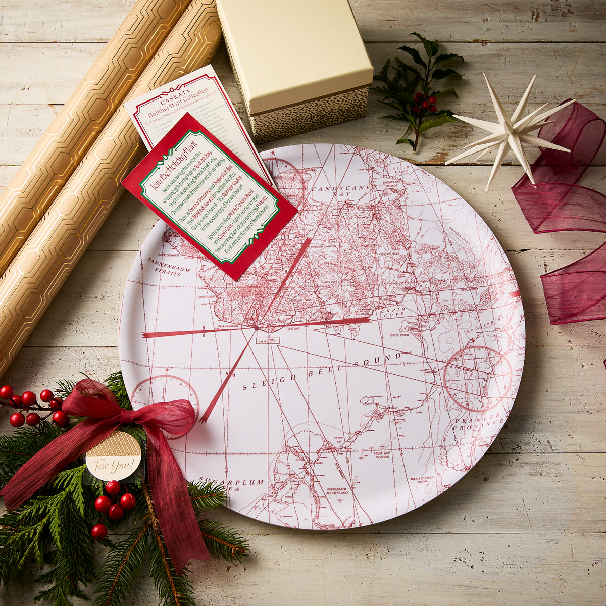 The Caskata White Holiday Hunt Large Round Tray sits on a wooden table with wrapped gifts, red ribbon, holiday cards, greenery, and holly—making it a sustainable and festive platter for your holiday gatherings.