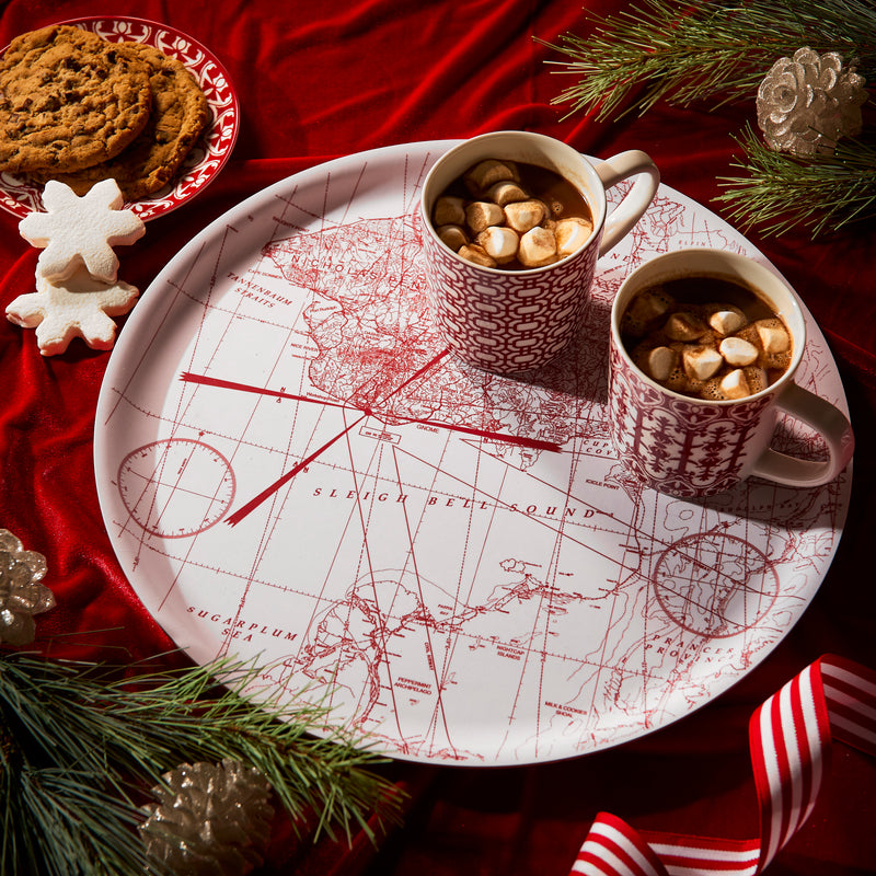 The Caskata White Holiday Hunt Large Round Tray features a red map design and holds two mugs of hot chocolate with marshmallows. Surrounded by cookies, pine branches, and holiday décor, it adds cheer while being a sustainable serving option.