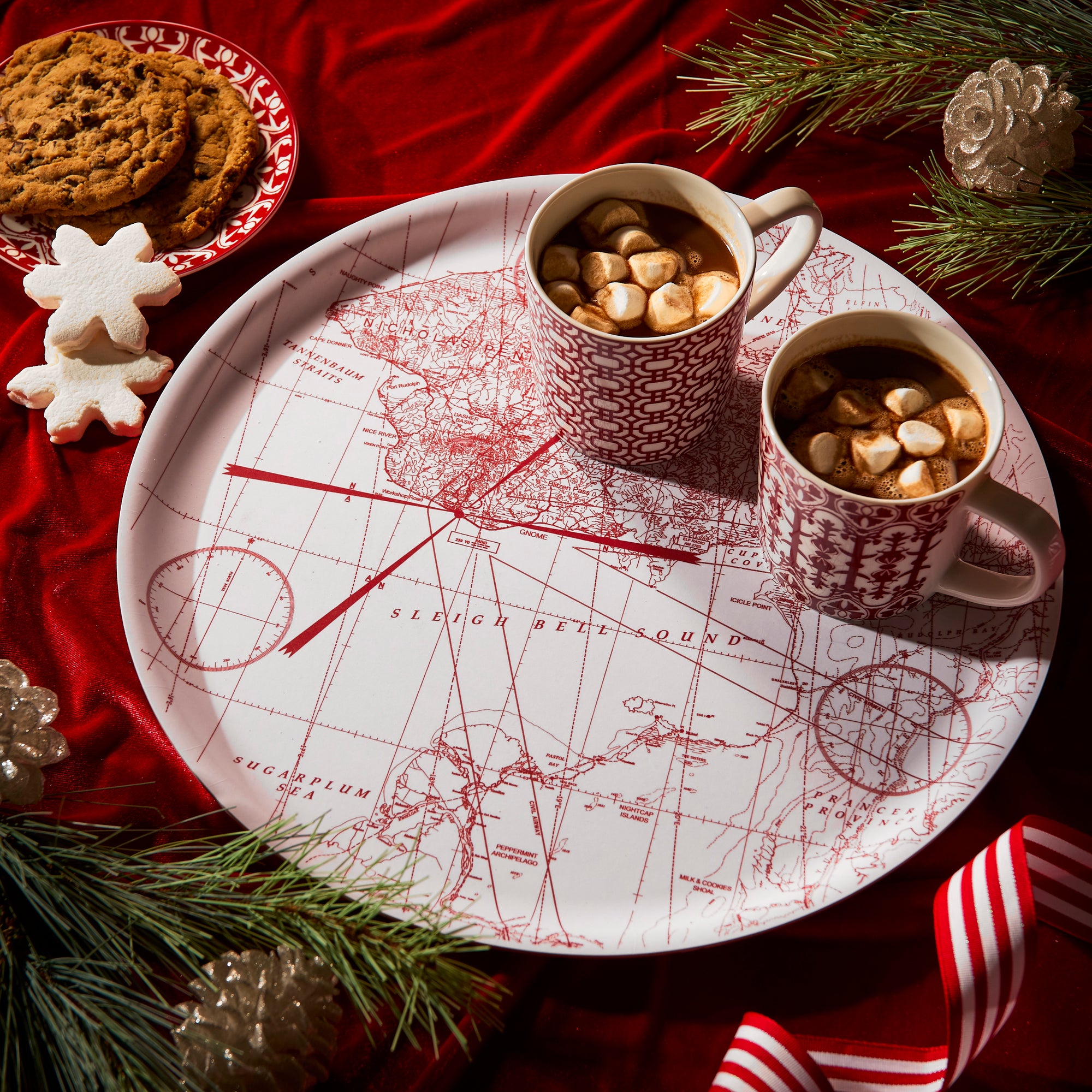 The Caskata "Holiday Cocoa" Bundle features four white porcelain mugs with red geometric patterns, placed in front of a birchwood serving tray adorned with a red vintage map design, all set against a white background.