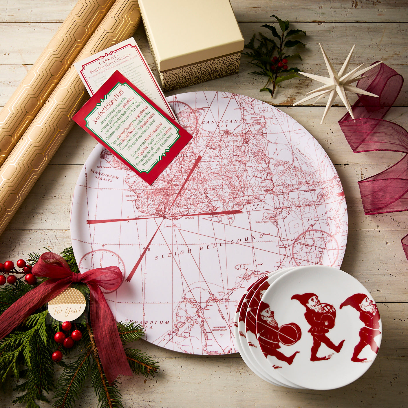 A festive flat lay showcases Caskata’s "Holiday Hunting" Bundle, with a decorative platter, Elves Dessert Plates, Santa bowls, gift wrap, boxes, ribbon, and holiday greenery on a wooden surface.