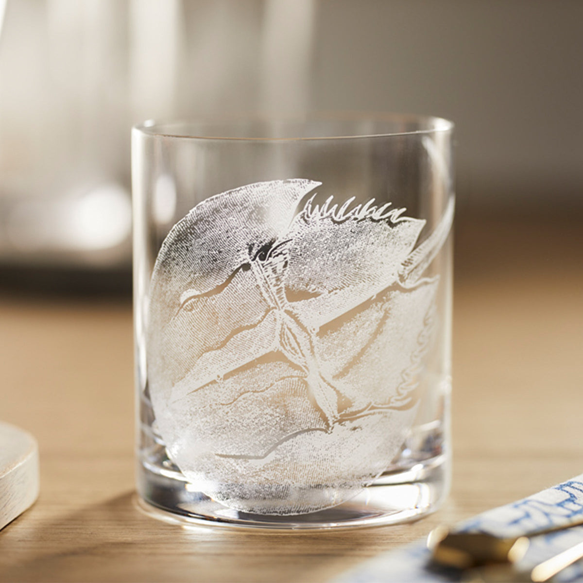 The Caskata Horseshoe Crab Rocks Glasses feature a frosted horseshoe crab design etched on clear glass with a light wooden surface.