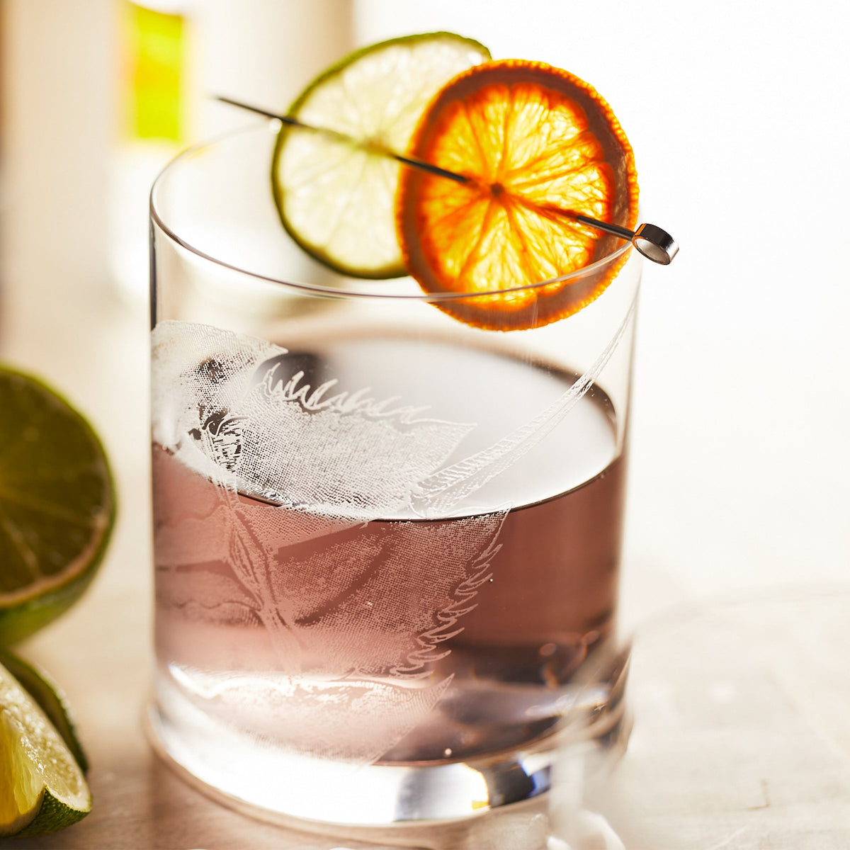 A glass of purple cocktail in a Caskata Horseshoe Crab Rocks Glass, adorned with lime and orange slices on a pick, sits beside lime wedges.