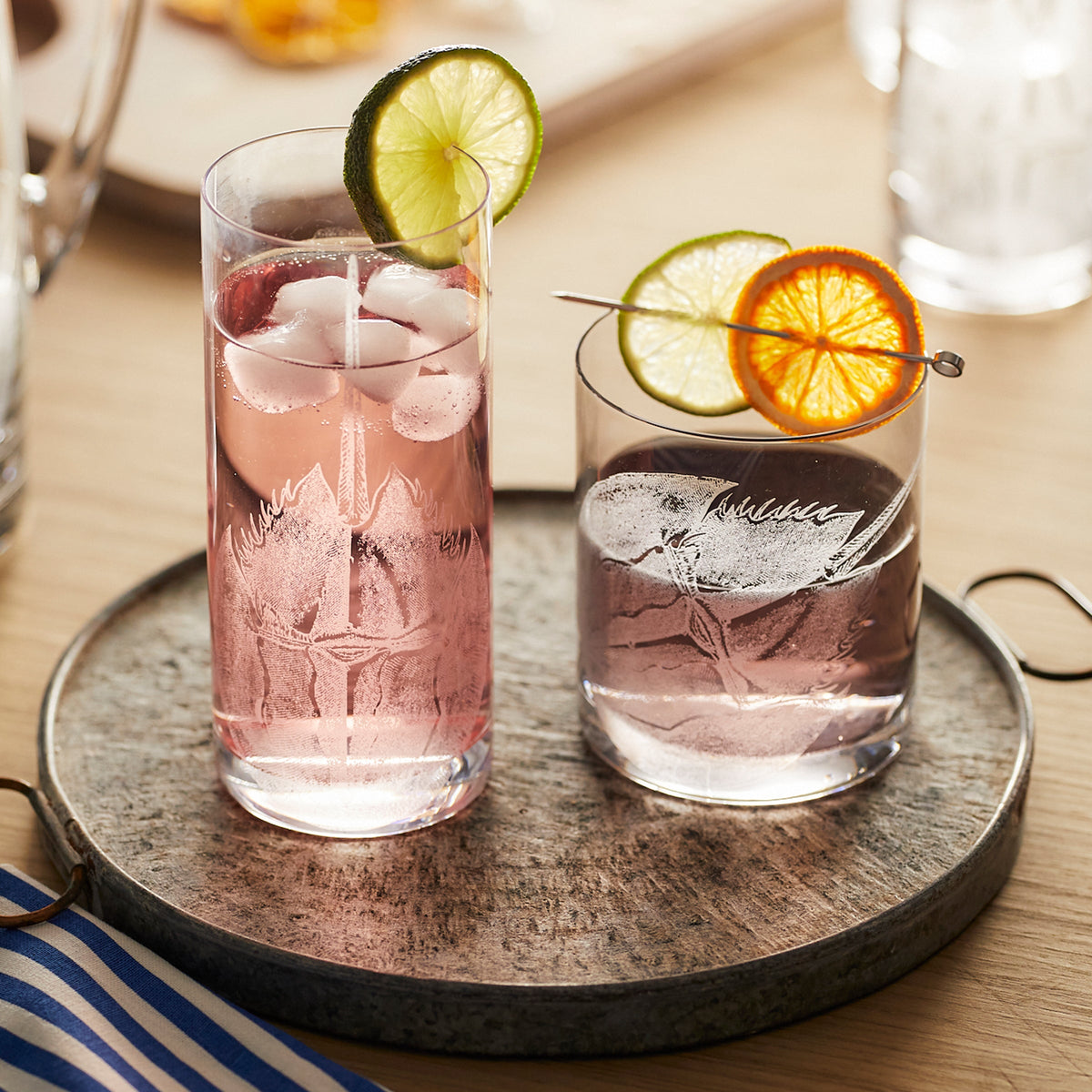 The Caskata Horseshoe Crab Rocks Glasses set includes two lead-free crystal glasses on a metal tray; the taller glass contains pink liquid with ice and a lime slice, while the shorter glass holds clear liquid with ice, accompanied by an artfully skewered lime and orange slice.