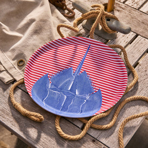 The Caskata Horseshoe Crab Large Round Birchwood Tray, featuring a blue horseshoe crab illustration, is crafted in Finland and pictured on a red and white striped plate with nautical accents nearby.