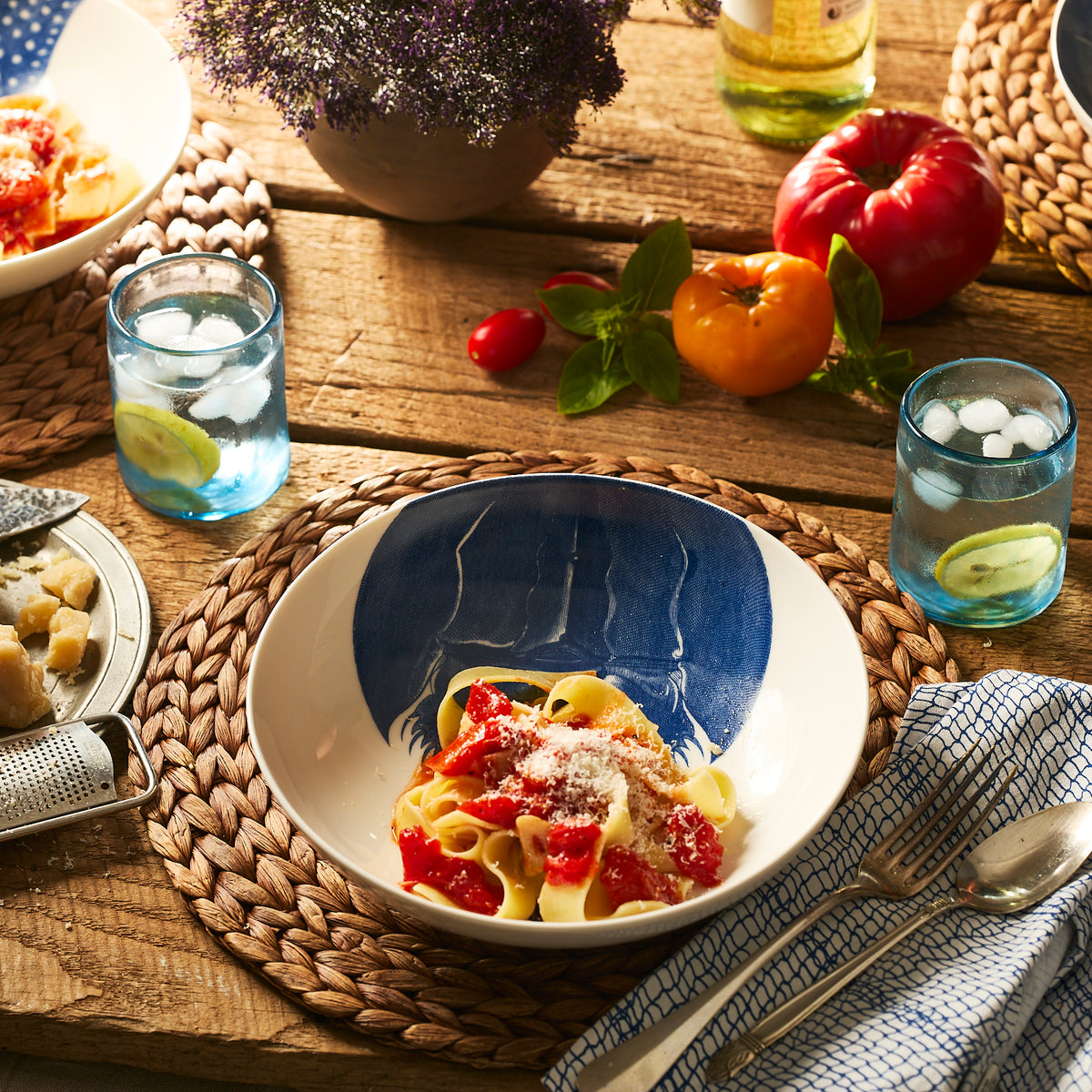 A Caskata Horseshoe Crab Entrée Bowl filled with pasta, tomato sauce, and Parmesan sits on a woven placemat beside two lime water glasses, fresh tomatoes, and a potted plant on a rustic wooden table.