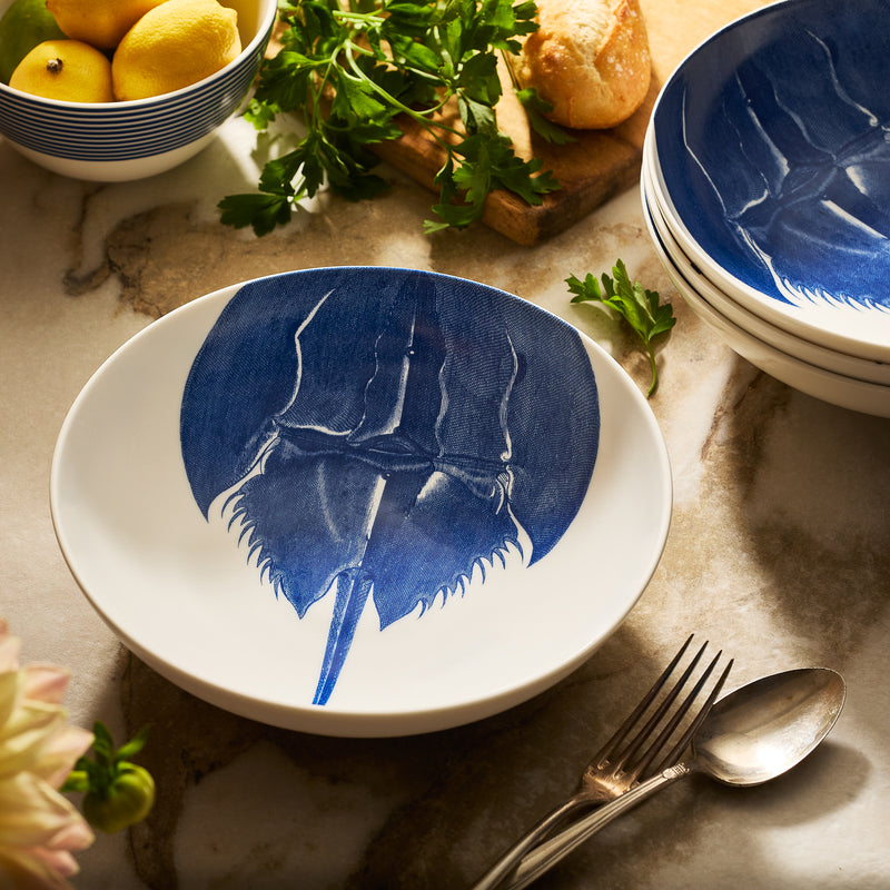 The Caskata Horseshoe Crab Entrée Bowl, crafted from white porcelain with a blue horseshoe crab design, rests on marble alongside silverware, stacked bowls, lemons, and parsley—capturing New England coastal charm.