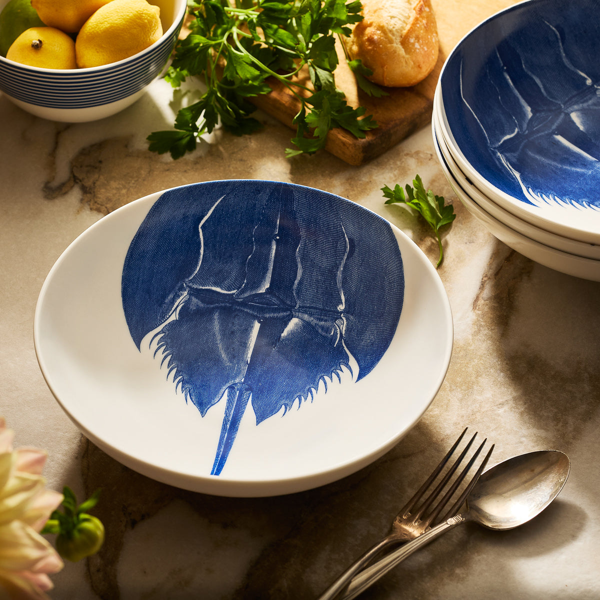 The Caskata Horseshoe Crab Entrée Bowl, crafted from white porcelain with a blue horseshoe crab design, rests on marble alongside silverware, stacked bowls, lemons, and parsley—capturing New England coastal charm.