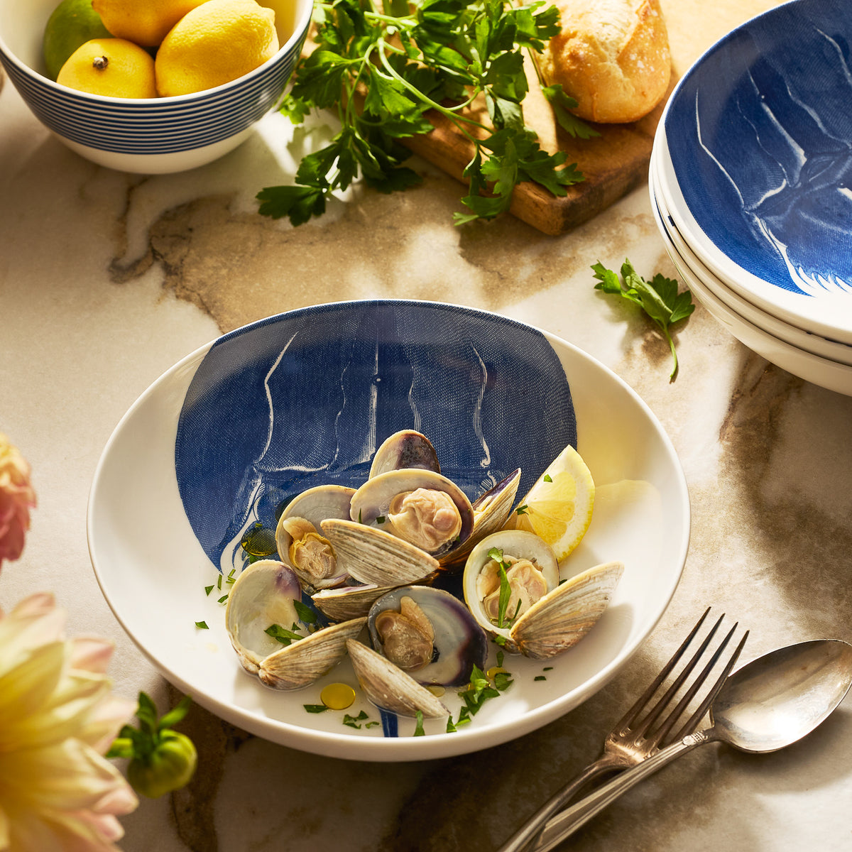 The Caskata Horseshoe Crab Entrée Bowl, filled with steamed clams and garnished with parsley and lemon, sits beside bread, utensils, and fresh herbs—capturing the classic elegance of New England dining.