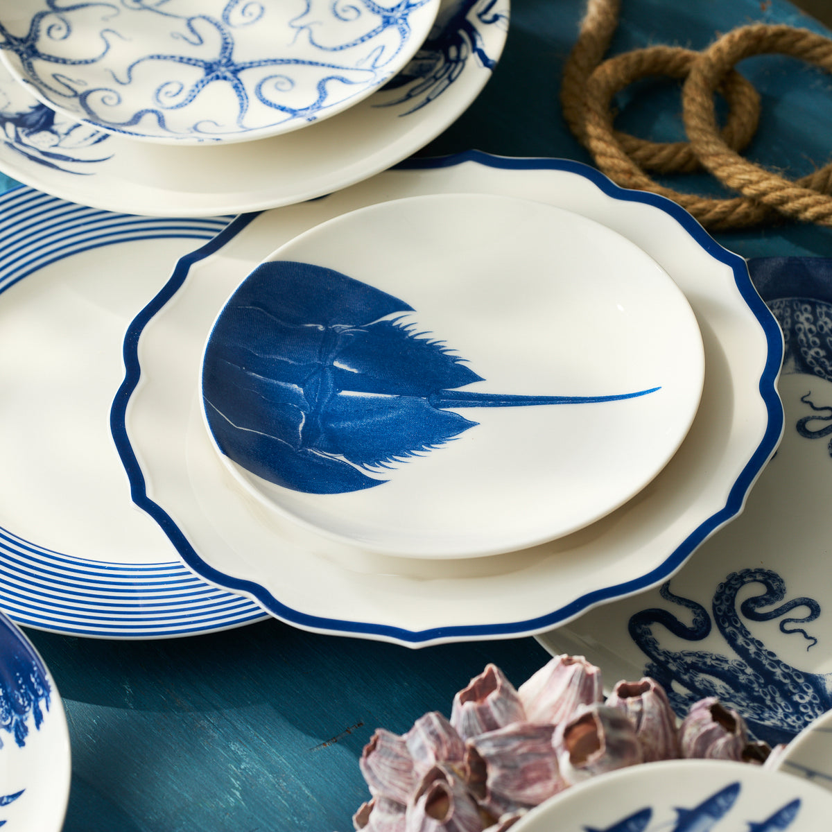 Caskata Artisanal Home’s Horseshoe Crab Coupe Salad Plate, featuring blue sea creature designs, is arranged with assorted contemporary dinnerware on a blue table accented by rope and barnacle decor.