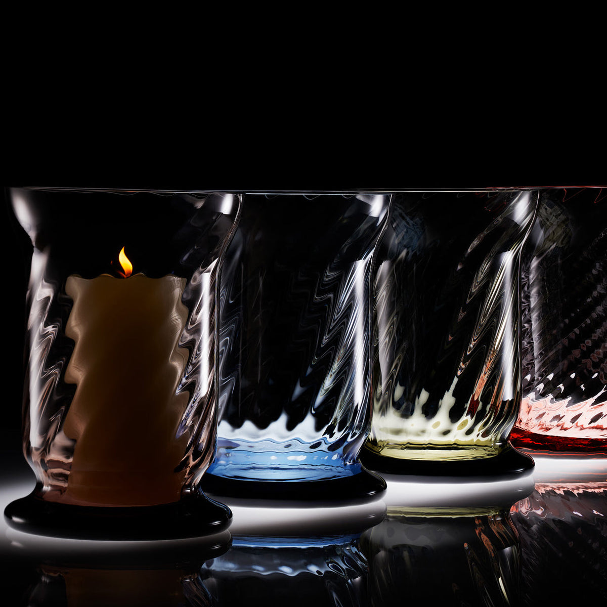 Row of multicolor Caskata Hurricanes with a moody dark background and one candle flickering in the foreground. Hurricanes are amber, ocean, smoke and rose colored.