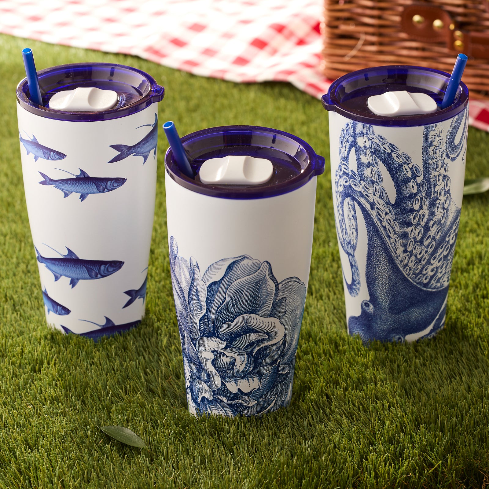 Three Peony Stainless Steel Insulated Tumblers by Caskata, each with distinct blue and white designs—fish, flower, and octopus—and matching blue straws, sit on the grass beside a picnic basket and a red checkered blanket. They are perfect for keeping your drinks cool in style during outdoor adventures.
