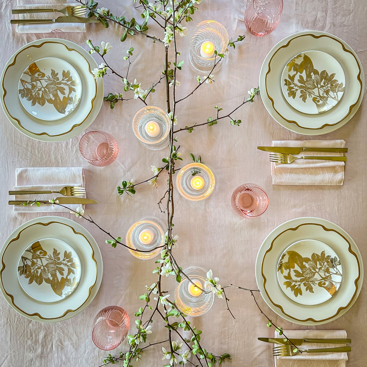 Bird&#39;s eye view of a spring tabletop featuring Caskata&#39;s Gold Arbor canapé plates on Grace Gold buffet plates around an assortment of Quinn mouth-blown glassware and a flowering branch centerpiece. 