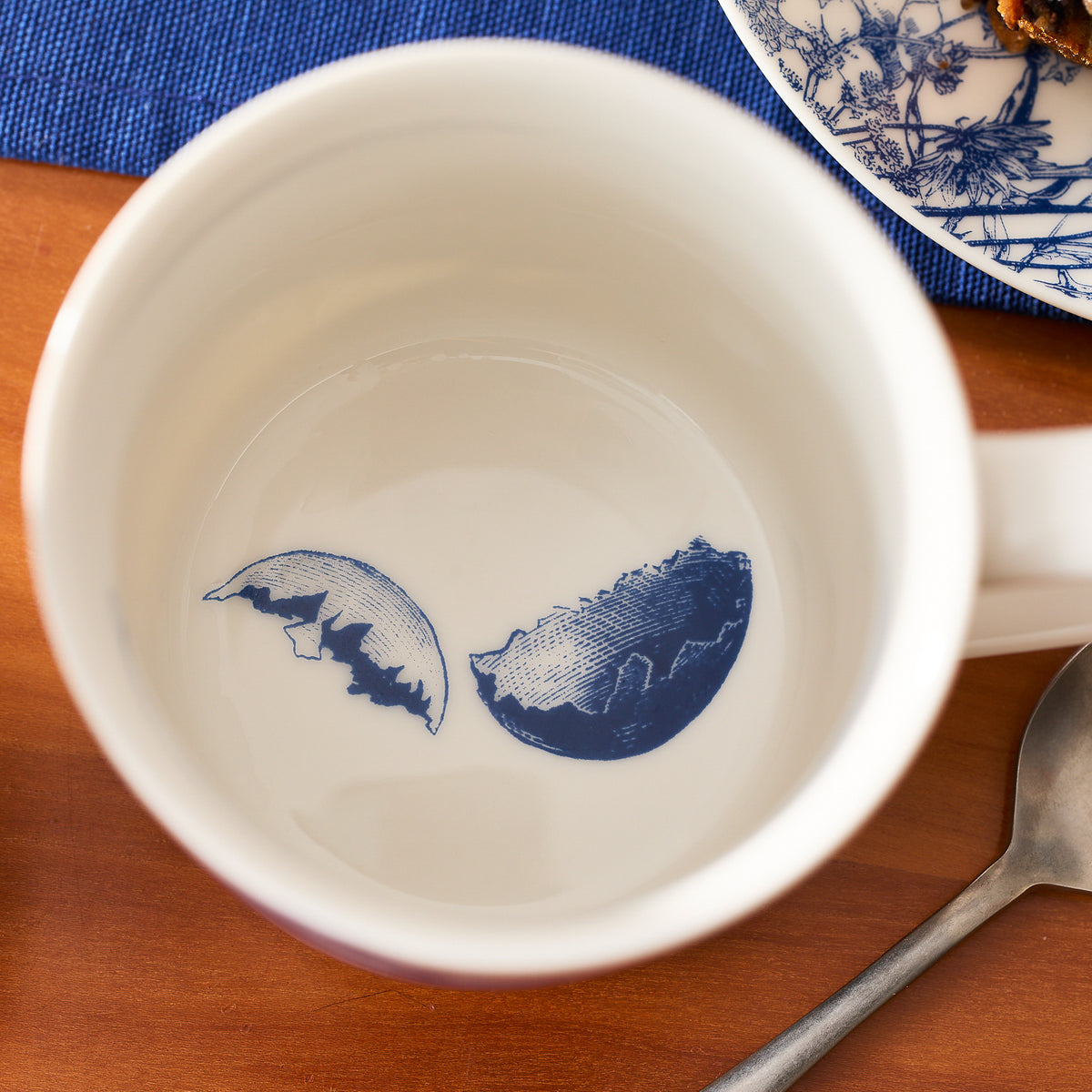 A Caskata Grumpy Chick Mug rests on a wooden table, paired with a spoon and a blue patterned plate.