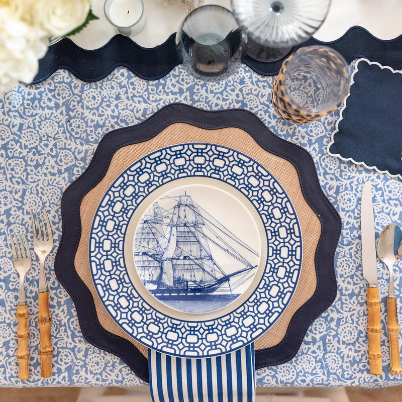 A dining table setting featuring heirloom-quality Rigging Small Plates by Caskata Artisanal Home, showcasing a blue and white patterned design with a ship illustration, placed on a navy scalloped placemat and blue printed tablecloth, accompanied by bamboo-handled cutlery, celebrating nautical heritage.