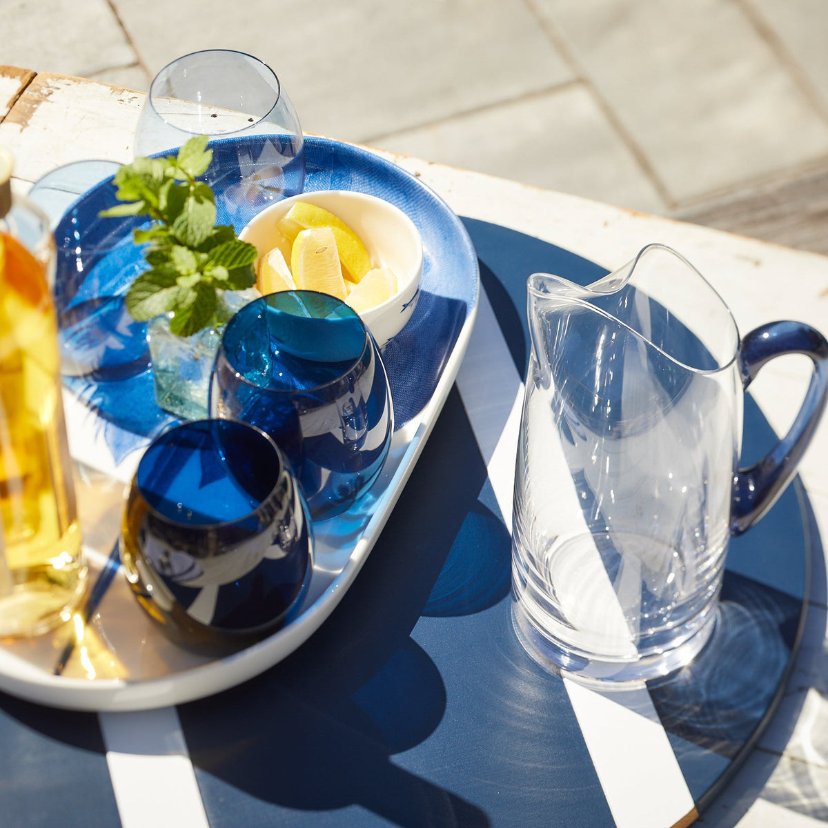 A Les Nuages Blue Handle Small Pitcher by Caskata and lead-free crystal glasses on a blue and white tray, accompanied by fresh lemons.