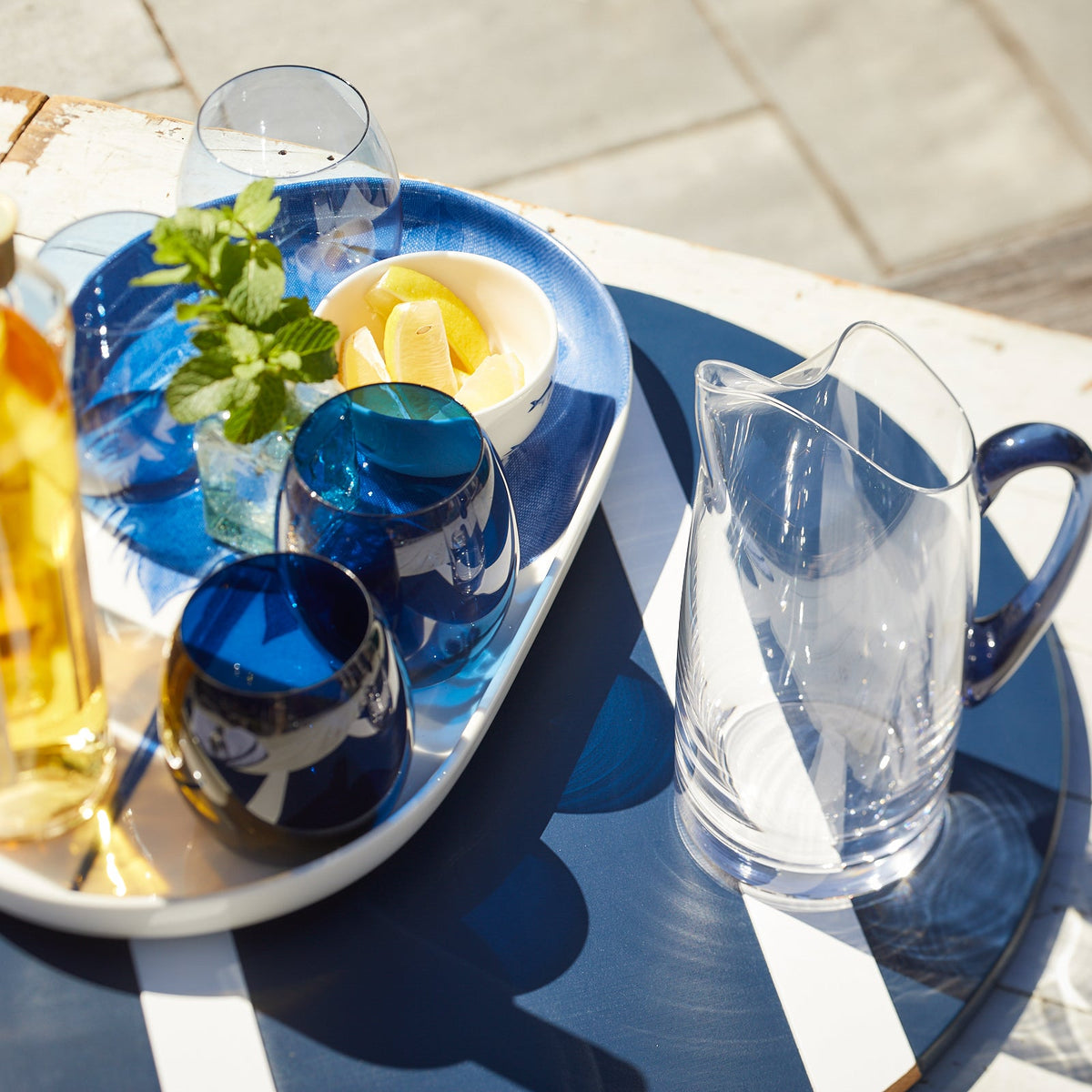 The Santorini Set Bundle by Caskata features Les Nuages Tumblers, a clear lead-free crystal pitcher, and stylish outdoor essentials on a blue and white striped table.