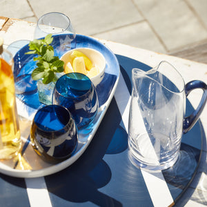 The Santorini Set Bundle by Caskata features Les Nuages Tumblers, a clear lead-free crystal pitcher, and stylish outdoor essentials on a blue and white striped table.