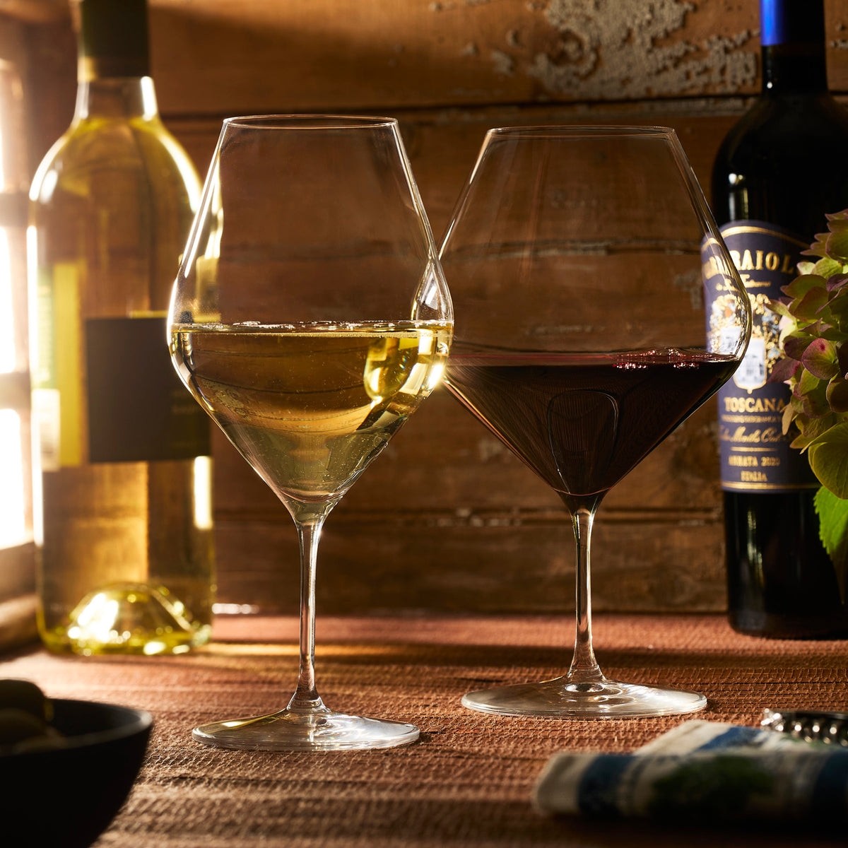 Two Caskata Library White Wine Glasses from the set of 4 are set on a table, filled with white and red wine, with two Library Collection wine bottles and a small flower arrangement in the background.